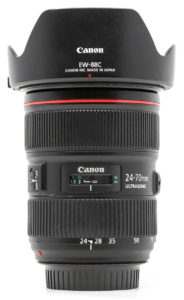 - Location de Studio équipé pour la Photo et la Vidéo Un objectif zoom Canon 24-70mm avec un pare-soleil, posé debout sur un fond blanc uni. L'objectif possède un stabilisateur d'image et une technologie ultrasonique, avec des indications de longueur focale visibles.